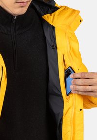 Żółta kurtka puchowa częściowo rozpięta, odsłania czarny sweter pod spodem. Niebieski smartfon jest wkładany do zapinanej wewnętrznej kieszeni.