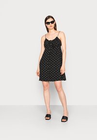 Vestido preto sem mangas com padrão floral, decote franzido e detalhes em botão. A modelo usa óculos de sol pretos e sandálias tipo slip-on.
