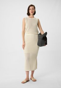 Knit two-piece outfit in cream featuring a sleeveless top and a mid-length skirt with a side slit, paired with neutral slip-on sandals and a striped bag.