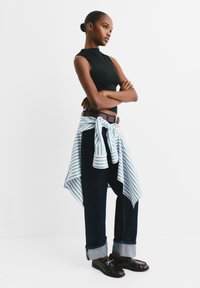 Schwarzes ärmelloses Top, dunkle Jeans mit hochgekrempelten Säumen, weißes und grünes gestreiftes Hemd um die Taille gebunden, brauer Gürtel, schwarze Slipper.