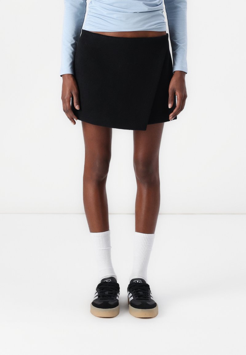 Jupe enveloppante noire en tissu doux, avec un design simple et des lignes épurées. Portée avec des baskets noires et des chaussettes blanches mi-mollet.