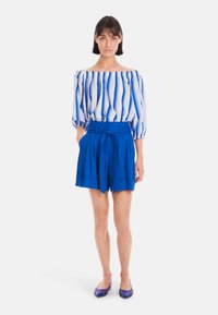 Schulterfreies Oberteil mit blauen und weißen vertikalen Streifen, kombiniert mit hochgeschnittenen blauen Shorts, die einen breiten Gürtel und Falten aufweisen.