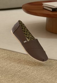 Braune Slip-On-Leinenschuhe mit innen einem Zickzackmuster, platziert über einem gewebten Teppich und in der Nähe eines runden Holztisches mit gestapelten Büchern.