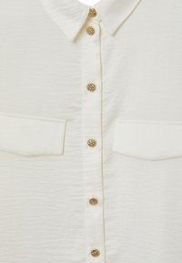 Camicia bianca a maniche lunghe con finitura texturizzata, caratterizzata da un colletto classico, bottoni decorativi dorati e due tasche sul petto.