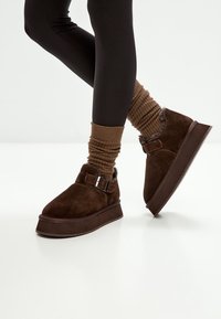 Legs wearing black leggings, brown slouchy socks, and dark brown suede platform shoes with buckle straps on a white floor.