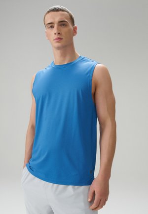 Jeune homme aux cheveux courts portant un débardeur de sport bleu et un short blanc, debout les mains dans les poches devant un fond neutre.