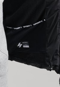 Intérieur de la veste noire avec doublure en mesh, bande avec logo "Superdry" et une étiquette montrant les détails de la marque et de la taille sur l'ourlet intérieur.