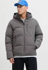 Młody mężczyzna w szarej pikowanej kurtce z kapturem i niebieskiej czapce, stojący z jedną ręką w kieszeni, patrzący w lewo na jednolitym tle.