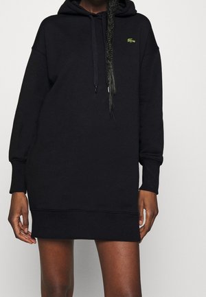 Person trägt ein schwarzes langärmeliges Kapuzenkleid mit einem kleinen grünen Krokodil-Logo auf der Brust und steht vor einem einfarbigen Hintergrund.