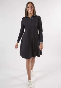 Robe noire à manches longues, col et patte de boutonnage. Rayures d'accent sur les poignets. Design de jupe évasée, associée à des baskets blanches.
