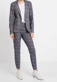 Grijze geruite blazer gecombineerd met een bijpassende tapered broek en een wit T-shirt. De stof lijkt soepel met een lichte textuur. Witte enkellaarsjes.