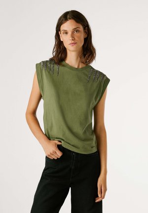 Camicia a maniche corte verde oliva con collo a giro, decorata con ornamenti argento sulle spalle e vestibilità comoda. Abbinata a pantaloni neri.