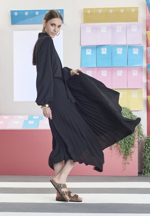 Black pleated dress with long sleeves and a cinched waist, paired with leopard print slides. Background features colorful paper displays.