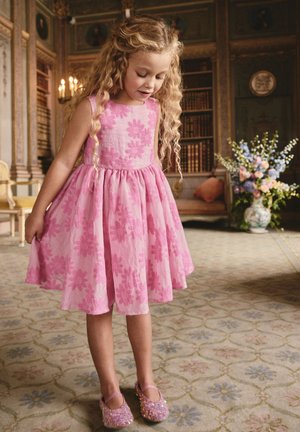 Junges Mädchen mit lockigem blondem Haar, das ein rosa geblümtes Kleid und glitzernde rosa Schuhe trägt, steht in einem kunstvoll dekorierten Raum mit Bücherregalen und Blumen.