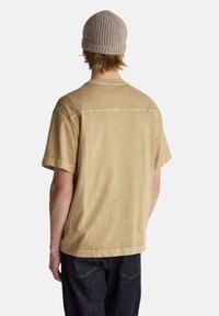 Béžové tričko s krátkým rukávem, jemnou texturou a kulatým výstřihem, doplněné tmavými džíny a lehkou pletenou čepicí. Minimalistický design.