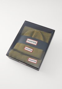 Zeleni pleteni kapica i šal u crnoj kutiji, sa crvenim i bijelim oznakama "HUNTER". Tkanina ima rebrastu teksturu.