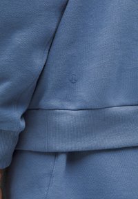 Jasnoniebieska bluza z ribbingu na dole i rękawach. Posiada małe wytłoczone logo. Materiał wydaje się miękki o gładkiej fakturze.