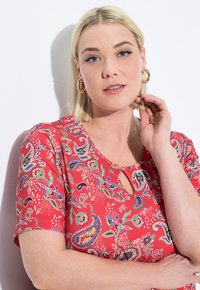 Červený top s krátkým rukávem, který má detail ve tvaru klíčové dírky a barevné vzory paisley. Hladká tkanina zobrazuje živé květinové akcenty.