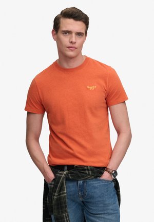 Junger Mann trägt ein oranges Superdry-T-Shirt, blaue Jeans und ein kariertes Hemd um die Taille gebunden, Hände in den Taschen, neutraler Hintergrund.
