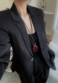 Svart blazer med strukturerade axlar och en enkel knappknäppning, kombinerad med en svart topp och ett mörkrött hänge på en snodd.