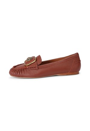 Bruine leren platte mocassin met geplooide neus en gouden decoratieve gesp bovenop, zijaanzicht tegen een witte achtergrond.
