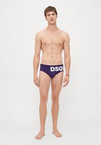 Jeune homme debout pieds nus, portant un maillot de bain violet foncé avec de grandes lettres blanches "DSQ" à l'avant, sur un fond blanc uni.