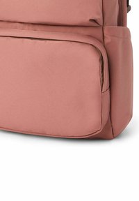 Mochila de tela rosa con una textura suave, que cuenta con un bolsillo frontal y un panel trasero acolchado para mayor comodidad. Diseño minimalista con acentos prácticos.