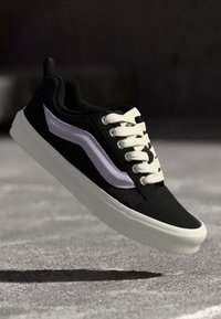 Svart joggesko med hvite snører og en teksturert design, som har et lilla stripeelement. Gummisåle og strømlinjeformet form.