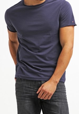 Homme portant un t-shirt uni bleu marine à manches courtes et un jean foncé, debout avec une main dans la poche et l'autre reposant près de la taille.