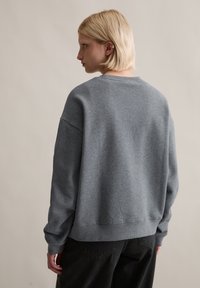 Szary sweatshirt z okrągłym dekoltem, z opuszczonymi ramionami i ściągaczami. Miękka tkanina o luźnym kroju, pokazany z tyłu na neutralnym tle.