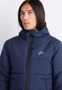 Granatowa kurtka puchowa Nike z zapięciem na zamek, regulowanym kapturem i logo na klatce piersiowej. Posiada pikowaną strukturę oraz przednie kieszenie.