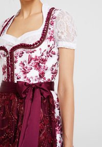 Bloemendess met een witte lijfje, bordeauxrode riem in de taille en intricaten kantensleeven. De rok heeft gedetailleerde borduursels en kralen.