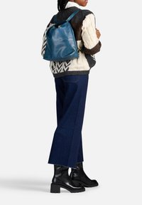 Zaino in pelle blu con una forma strutturata, spallacci regolabili e dettagli in oro. Indossato sopra un maglione di maglia e jeans.