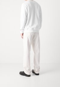 Sudadera blanca con puños acanalados, combinada con pantalones sueltos de color claro y calzado negro, capturada desde atrás contra un fondo blanco.