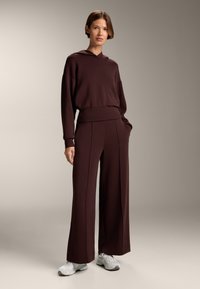 Bordowa bluza z kapturem o luźnym fasonie, w komplecie z szerokimi spodniami pasującymi w kolorze, z kontrastowymi szwami i kieszeniami po bokach, zestawiona z białymi trampkami.