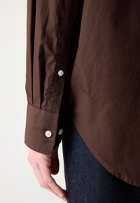 Gros plan d'une personne portant une chemise marron à manches longues avec des boutons blancs et un jean bleu foncé, en se concentrant sur le poignet et le bas de la manche.