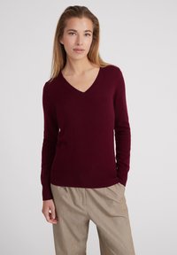 Burgunderfarbener V-Ausschnitt-Pullover aus weichem Stoff, mit langen Ärmeln und geripptem Saum, kombiniert mit hellen, plissierten Hosen.