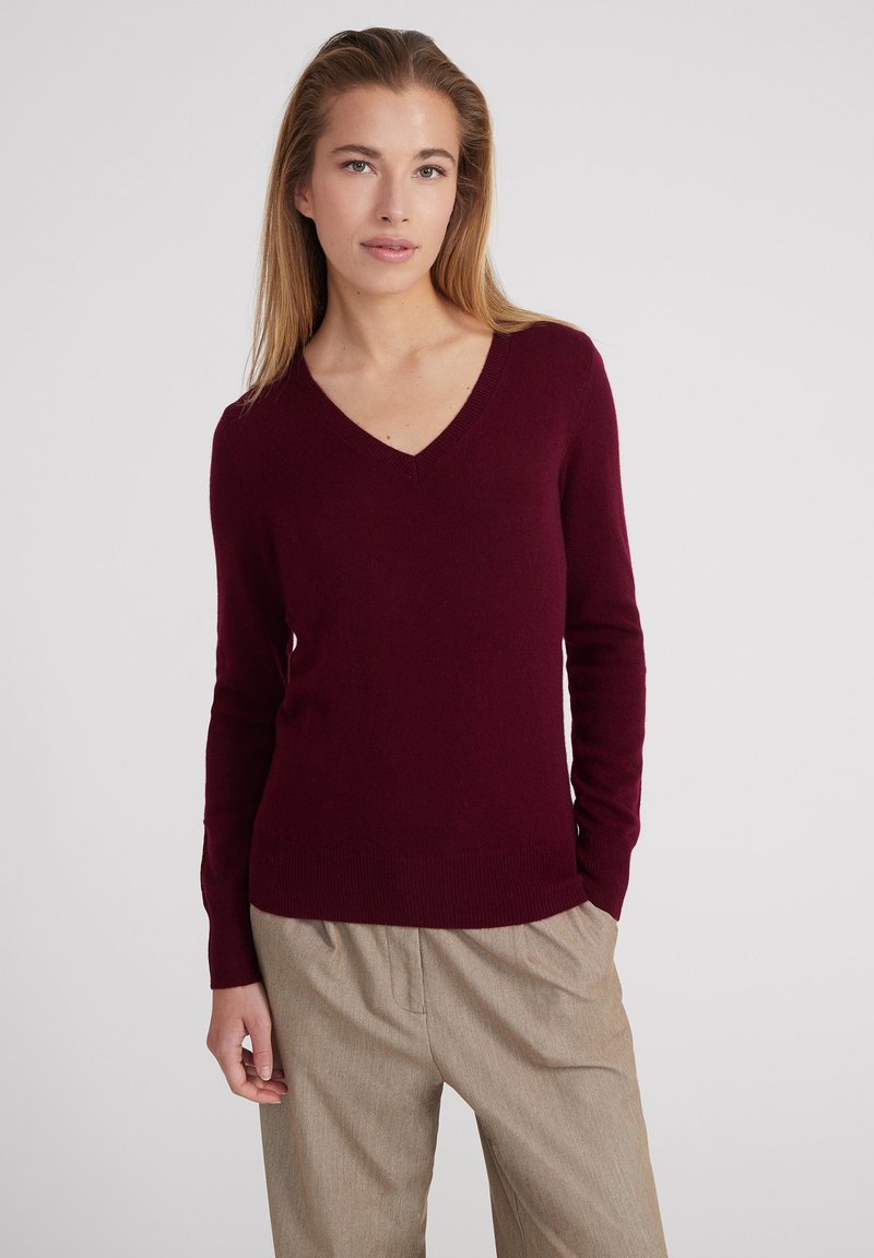 Burgunderfarbener V-Ausschnitt-Pullover aus weichem Stoff, mit langen Ärmeln und geripptem Saum, kombiniert mit hellen, plissierten Hosen.