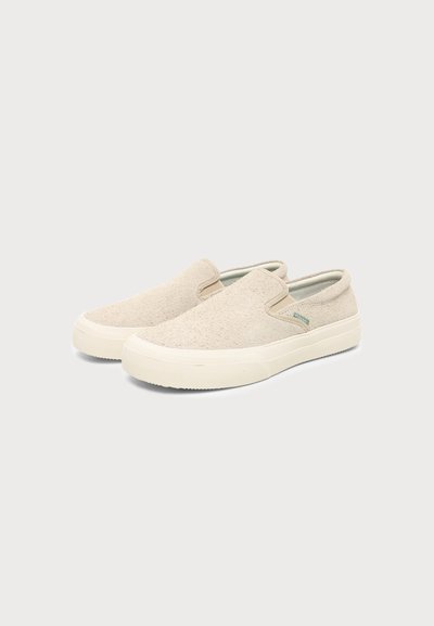 Ζευγάρι μπεζ sneakers τύπου slip-on με υφασμάτινο επάνω μέρος με υφή και λευκή λαστιχένια σόλα σε απλό ανοιχτό φόντο.