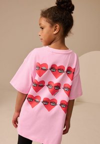 Rosa oversized t-shirt med hjärt- och ögondiagram på baksidan. Korta ärmar, mjukt tyg, avslappnad passform, idealisk för komfort och lek.