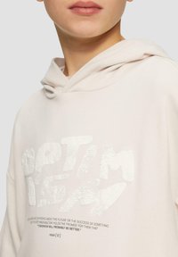 Lichtroze hoodie met witte textuur grafische tekst "OPTIMISME" en een citaat eronder. Voorzien van een trekkoordkap en zachte stof textuur.