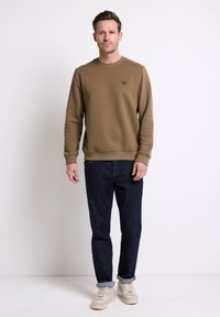 Felpa marrone con tessuto testurizzato, colletto a girocollo e piccolo logo sul petto, abbinata a jeans blu scuro e sneakers beige.