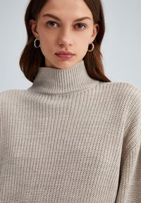 Beige gebreide coltrui met een textuurpatroon, voorzien van een hoge kraag en geribde manchetten. Zilveren ringoorbellen als accessoires.