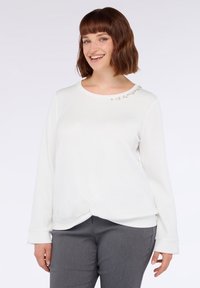 Maglietta bianca a maniche lunghe con scollo rotondo, caratterizzata da sottili decorazioni vicino al colletto e un dettaglio annodato all'orlo. Abbinata a pantaloni grigi.