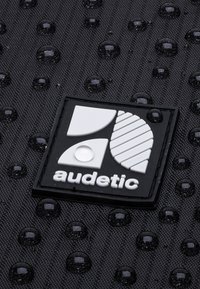 Superficie nera testurizzata con gocce d'acqua e un logo bianco con forme geometriche e la parola "audetic" in fondo.