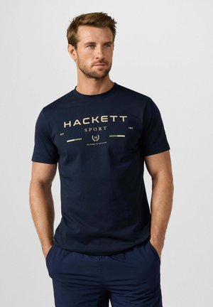 Man met kort bruin haar die een marineblauw Hackett Sport T-shirt en bijpassende broek draagt, staat met handen in de zakken en kijkt naar links.