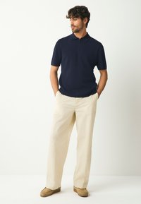 Rövid ujjú, galléros tengerészkék póló, krémszínű, bő szabású nadrággal és barna bebújós cipővel párosítva. Egyszerű dizájn.