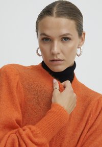 Oranžinė megzta suknelė su tekstūriniu paviršiumi, turinti sutrauktus rankogalius ir aukštą kaklą, dėvima ant juodos turtlenkės. Apvalūs pakabukai auskarai.