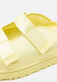Sandales en caoutchouc jaunes avec de larges sangles en tissu, présentant des détails texturés et un accent de marque. Semelle épaisse avec des motifs en rainure pour l'adhérence.