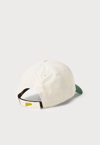 Baseball-lippalakki, jossa on kermaisen värinen puuvillapinta, vihreä lippa ja säädettävä remmi takana, jossa on keltainen logotunnus.